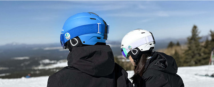 Two people wearing ski helmets on a snowy slope with trees and mountains in the background.